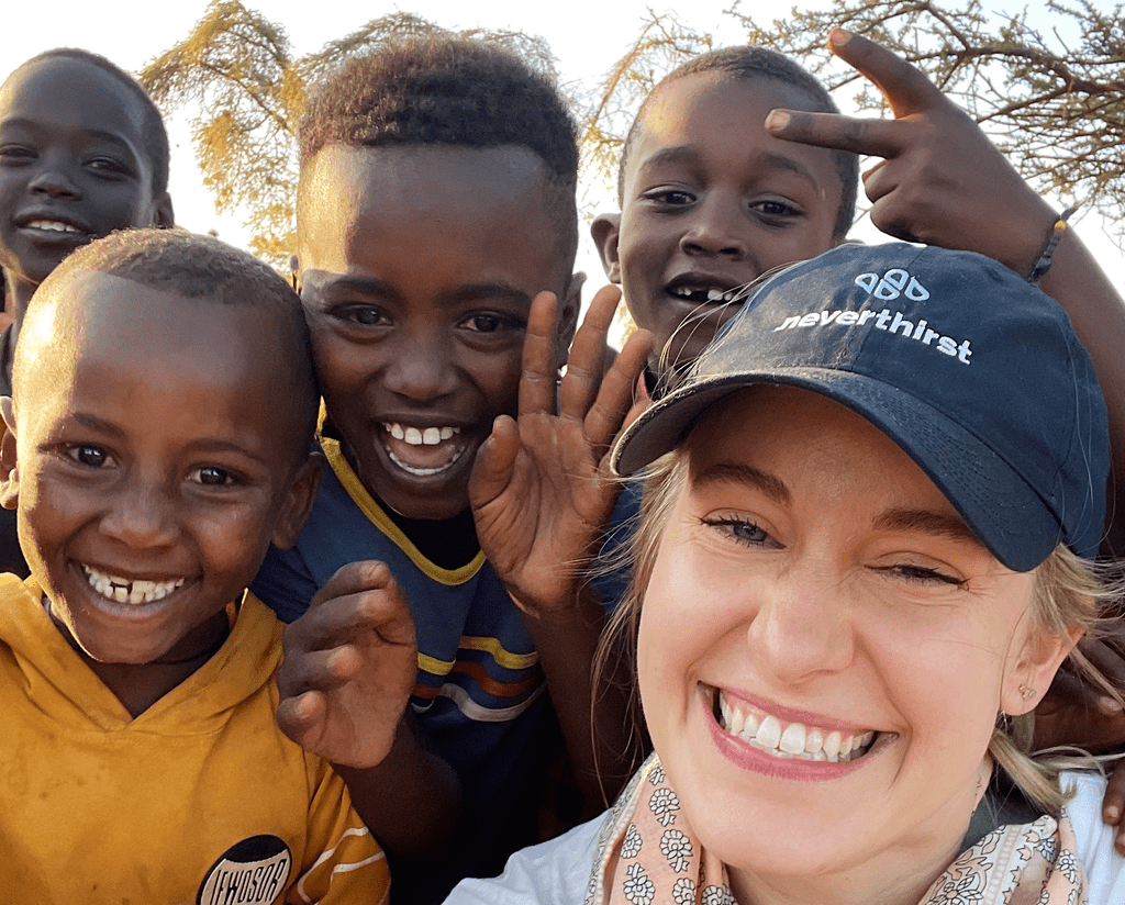 A group of smiling children gather around a woman in a cap who is also smiling and taking a selfie outdoors. The children are waving and appear cheerful, and the background shows trees and sunlight.
