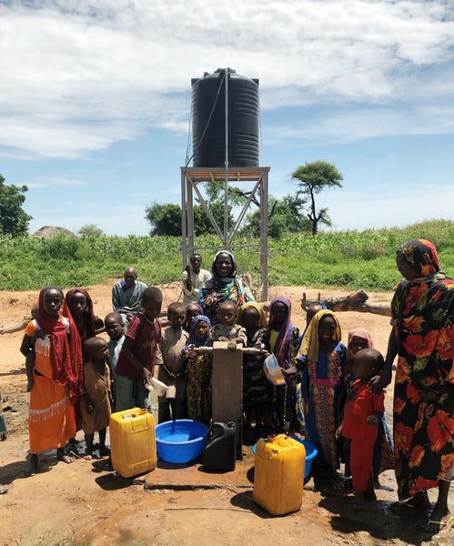 A group of children and adults gather around a water pump with containers, under a tank on a metal stand, in a rural outdoor setting with green vegetation and a blue sky.