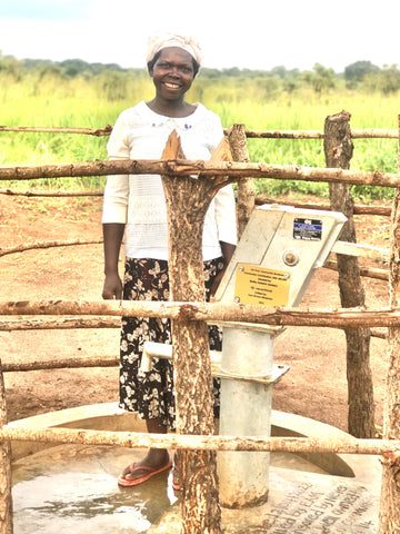 A woman in a white sweater, patterned skirt, and headscarf stands smiling by a hand water pump, surrounded by a rustic wooden fence in a rural outdoor setting with green fields in the background.