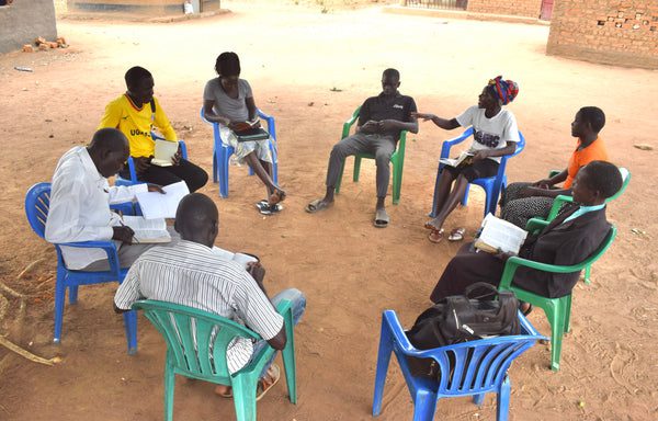 A group of eight people sit in a circle on plastic chairs outdoors, engaged in discussion and reading from books or papers, under open shade on sandy ground.
