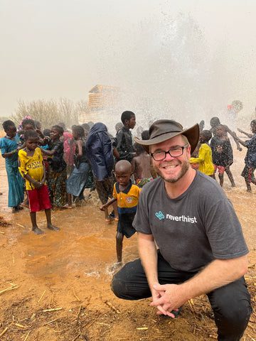 A smiling man in glasses and a hat kneels in the foreground wearing a Neverthirst shirt, while children play and splash in water behind him in a rural, outdoor setting.