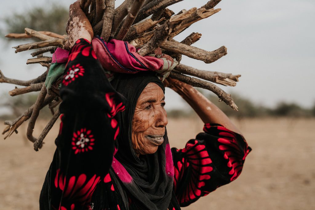 An elderly woman wearing a black and red patterned garment carries a bundle of firewood on her head in a dry, open landscape. She looks to the side, with one hand balancing the wood.
