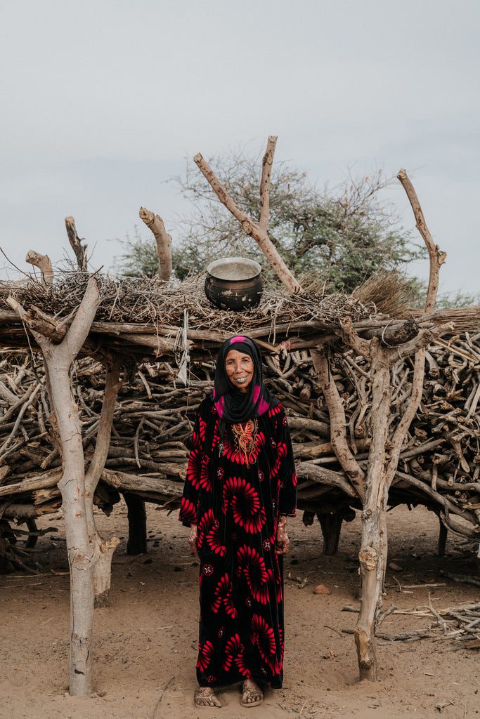 A woman in a black and red patterned dress stands smiling in front of a structure made from stacked branches and wood in a dry, sandy area with sparse trees.