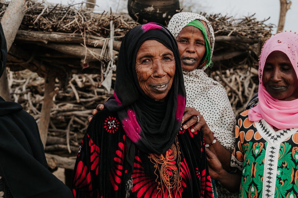 Three women in colorful clothing and headscarves stand together outdoors, smiling warmly. The background shows a rustic wooden structure and dry branches, suggesting a rural setting.