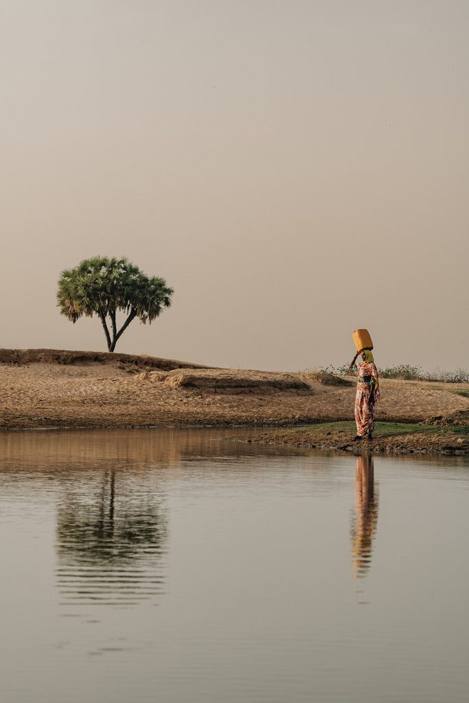 A person stands by a calm body of water carrying a yellow container on their head, with a single palm tree and sandy landscape in the background under a hazy sky.
