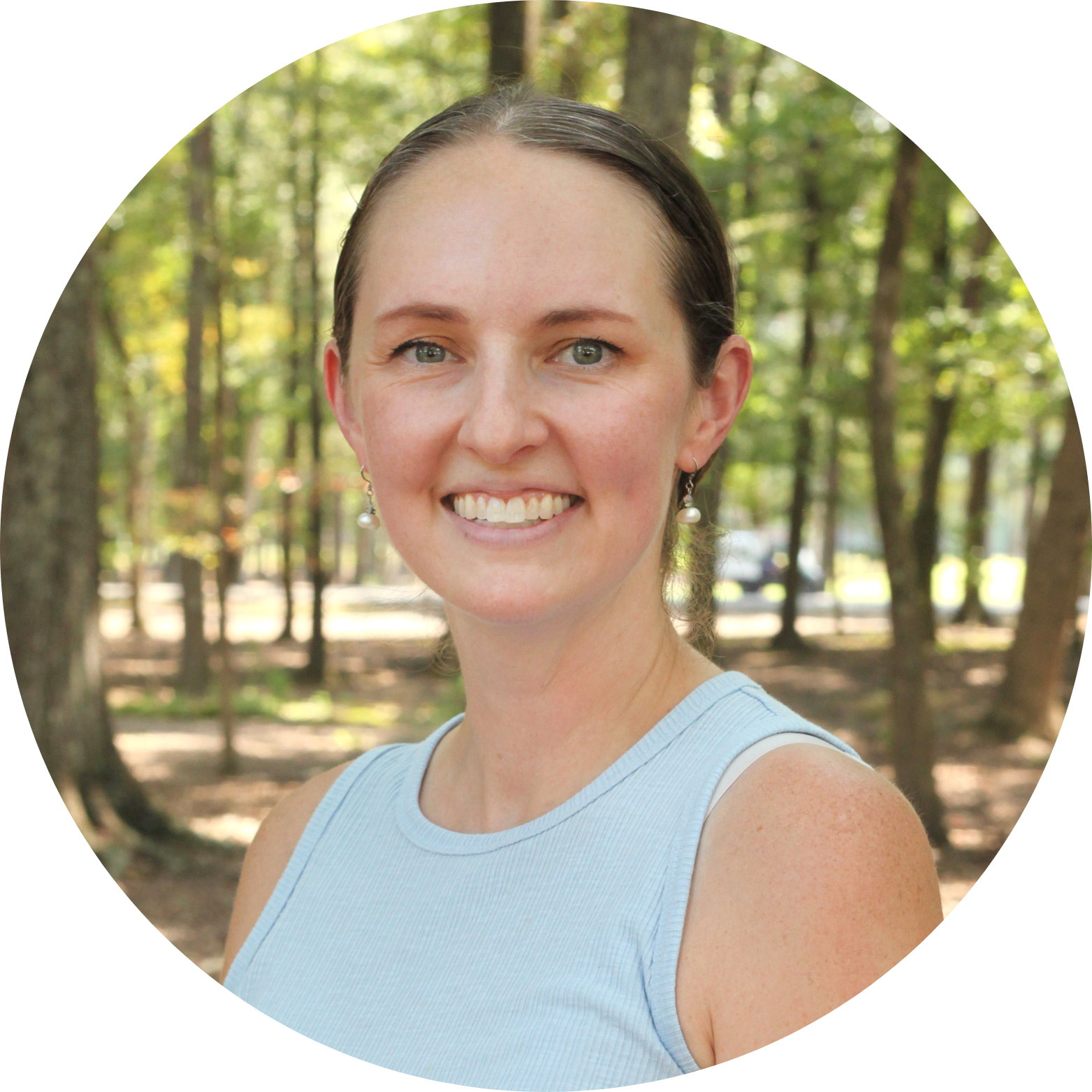 A woman with light skin and brown hair tied back smiles at the camera. She is wearing a light blue sleeveless top and earrings, standing outdoors in a wooded area with sunlight filtering through the trees.