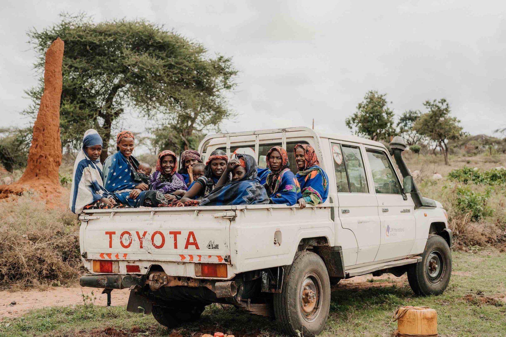 A group of women and children in colorful clothing sit in the back of a white Toyota pickup truck parked on a dirt path in a rural landscape with trees and a large termite mound.