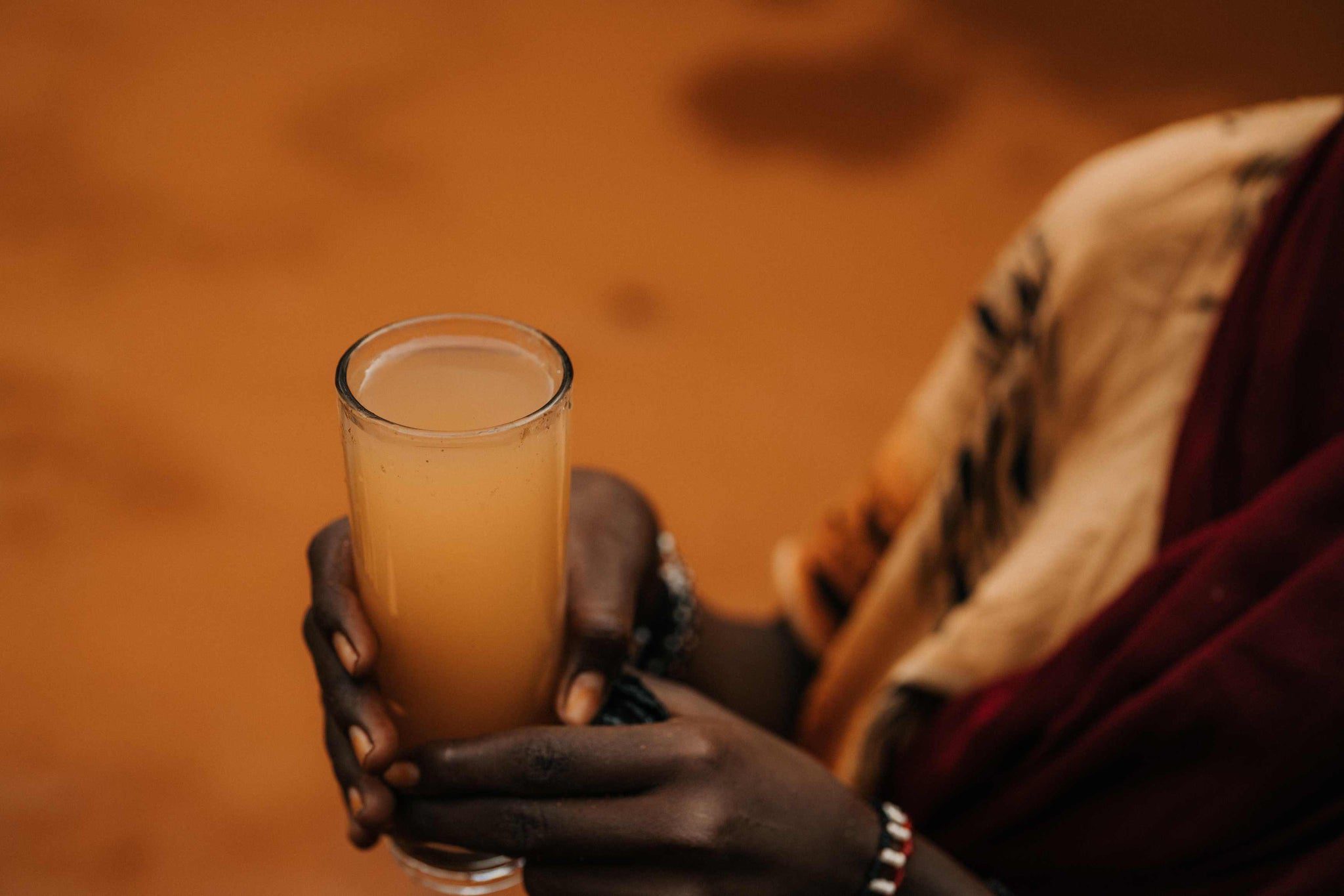 A person with dark skin holds a tall glass of cloudy, light brown liquid. The background is blurred and earthy brown, and part of the person’s patterned clothing and bracelets is visible.