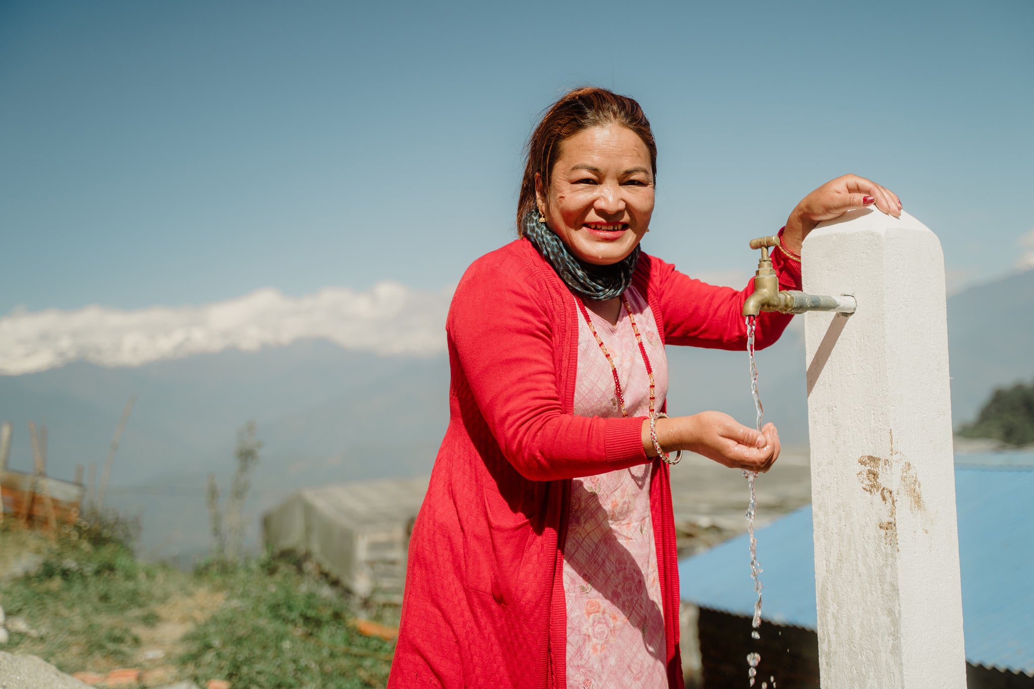 A woman in a red coat smiles while collecting water from an outdoor tap, with mountains and a clear sky in the background.
