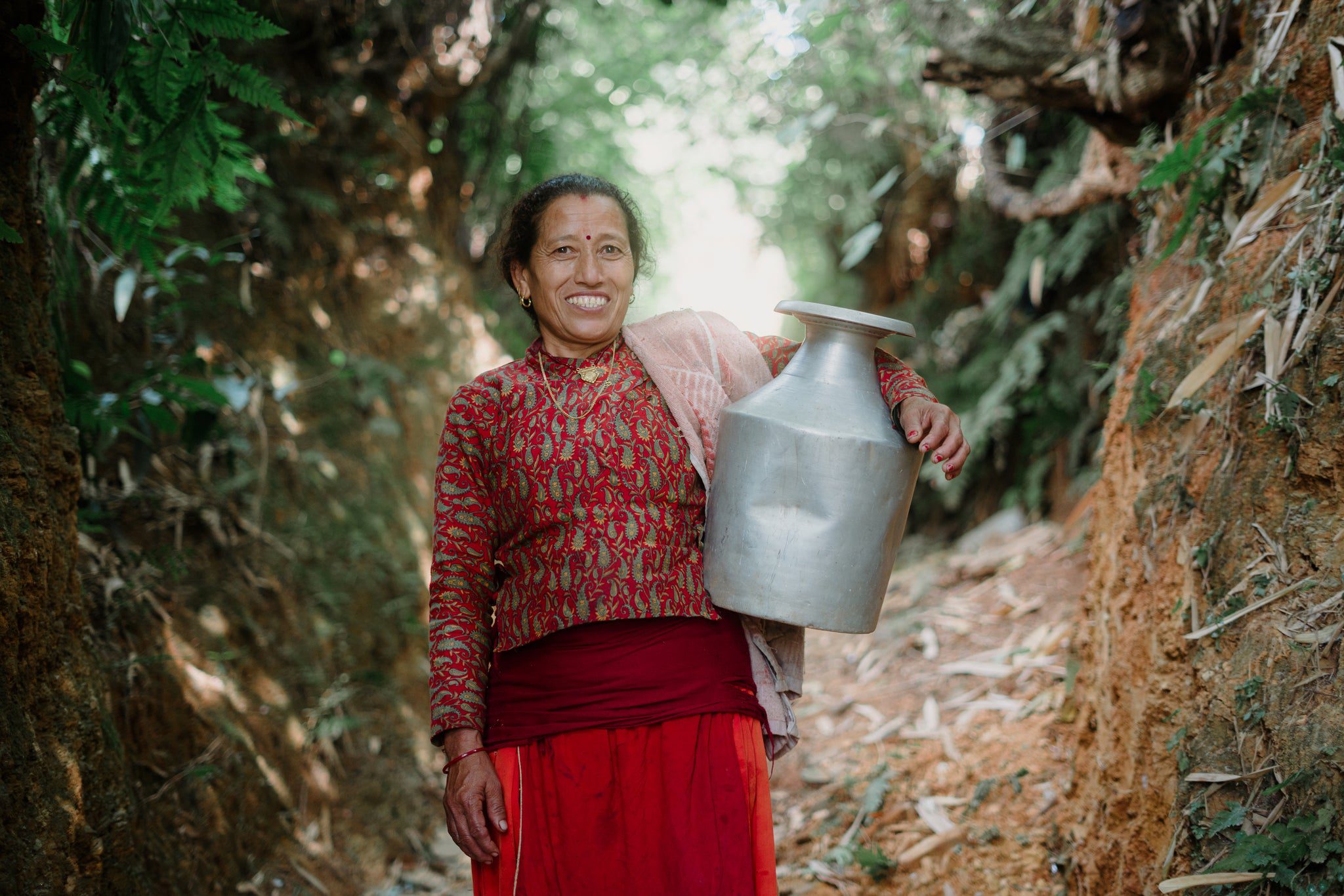 A woman in a red patterned top and skirt smiles while carrying a large metal container on her shoulder, standing on a dirt path surrounded by greenery.
