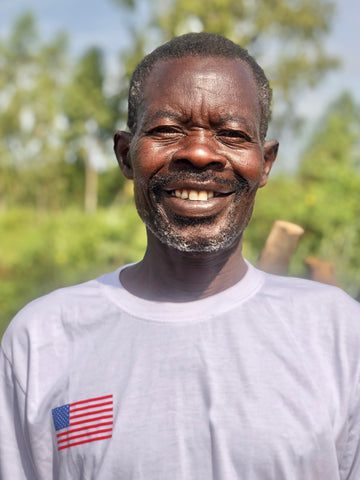 A smiling man with short hair and a beard stands outdoors wearing a white T-shirt with a small American flag patch on the chest. The background is blurred greenery and trees.
