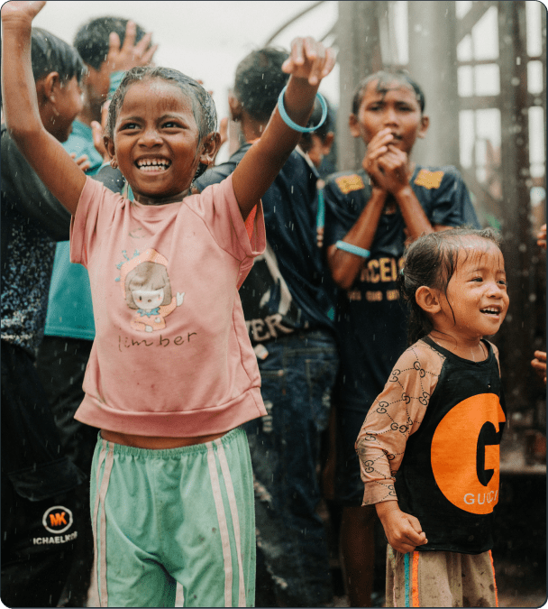 A group of smiling children play outside in the rain. The girl in front, wearing a pink shirt and green shorts, raises her arms joyfully, while other children laugh and watch in the background.