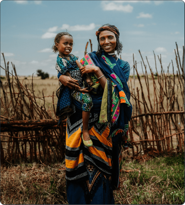 A woman dressed in colorful traditional clothing stands outdoors, smiling and holding a young girl. They are in front of a rustic wooden fence with a grassy field and blue sky in the background.