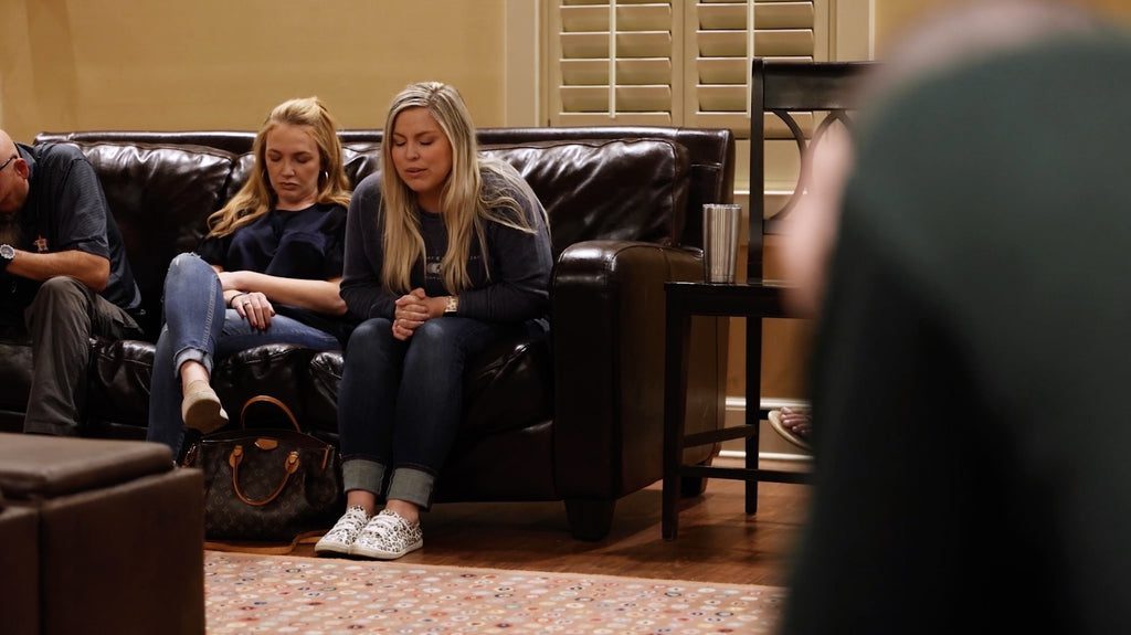 Three people sit on a dark couch indoors, two women in front with heads bowed and hands clasped, appearing to pray. A brown bag is on the floor near them. The room has pale walls and shutters.
