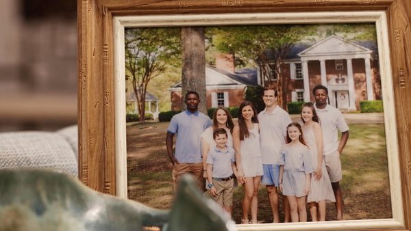 A framed photo shows a group of eight people, including adults and children, standing and smiling outdoors in front of a large brick house with columns and trees in the background.