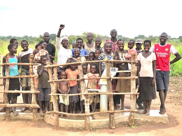 A group of children and adults in colorful clothing stand smiling and cheering around a wooden fence enclosing a water pump in a rural, grassy area.