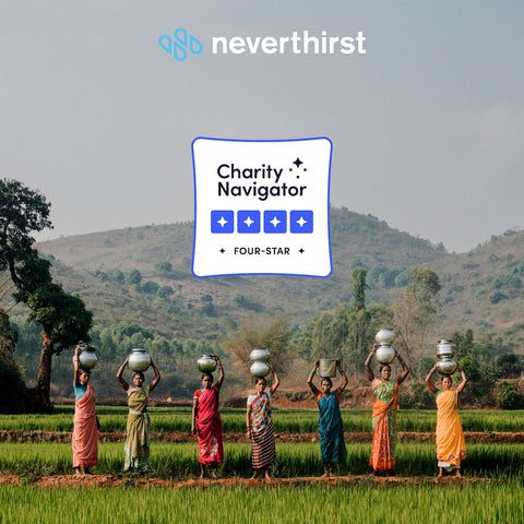 Seven women in colorful sarees stand in a green field balancing water pots on their heads. Hills and trees are in the background. The image features Neverthirst’s logo and a Charity Navigator four-star rating badge.