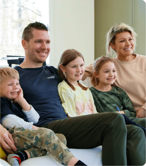 A smiling family of five sits together on a couch. An adult man and woman sit on either end, with three children between them, all appearing happy and relaxed in a brightly lit room.