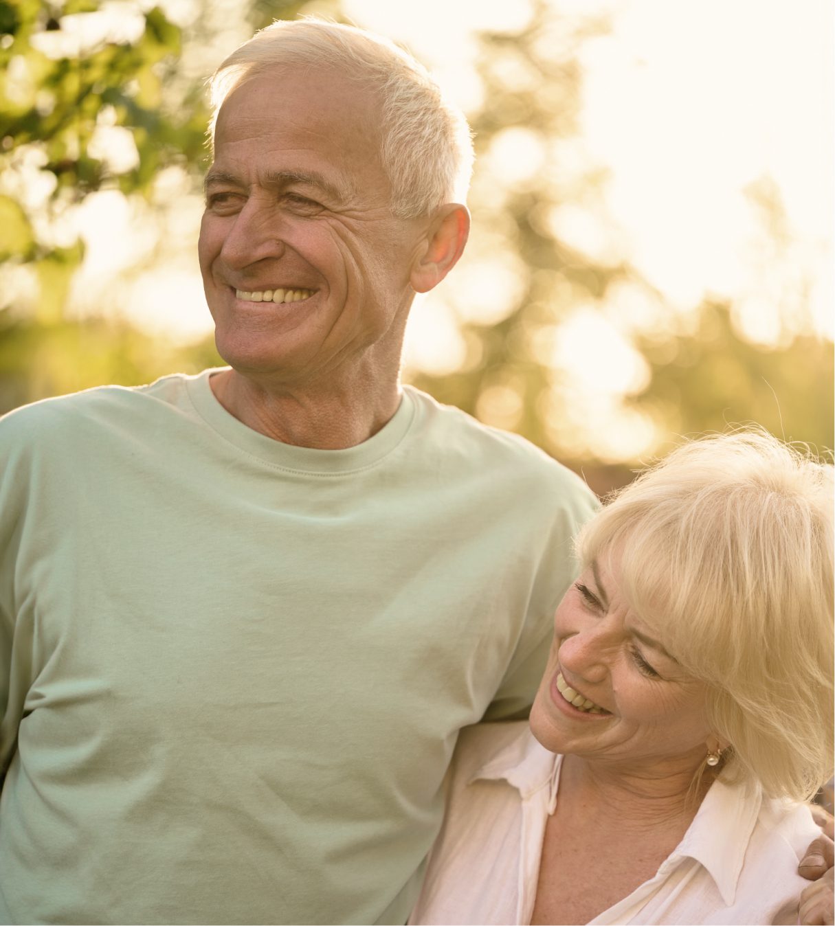 An older couple smiles warmly while standing outdoors in soft sunlight. The man has his arm around the woman, who leans affectionately on his shoulder. Both appear happy and relaxed, with greenery in the background.