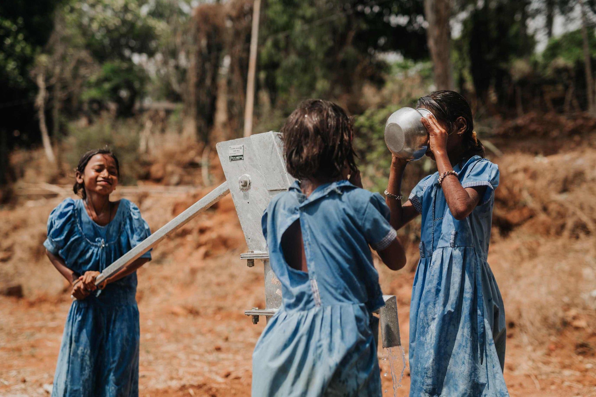 Three girls in blue dresses use a hand pump to draw water outdoors. One girl pumps, another drinks from a metal bowl, and the third girl smiles nearby. The setting is rural with dry grass and trees in the background.