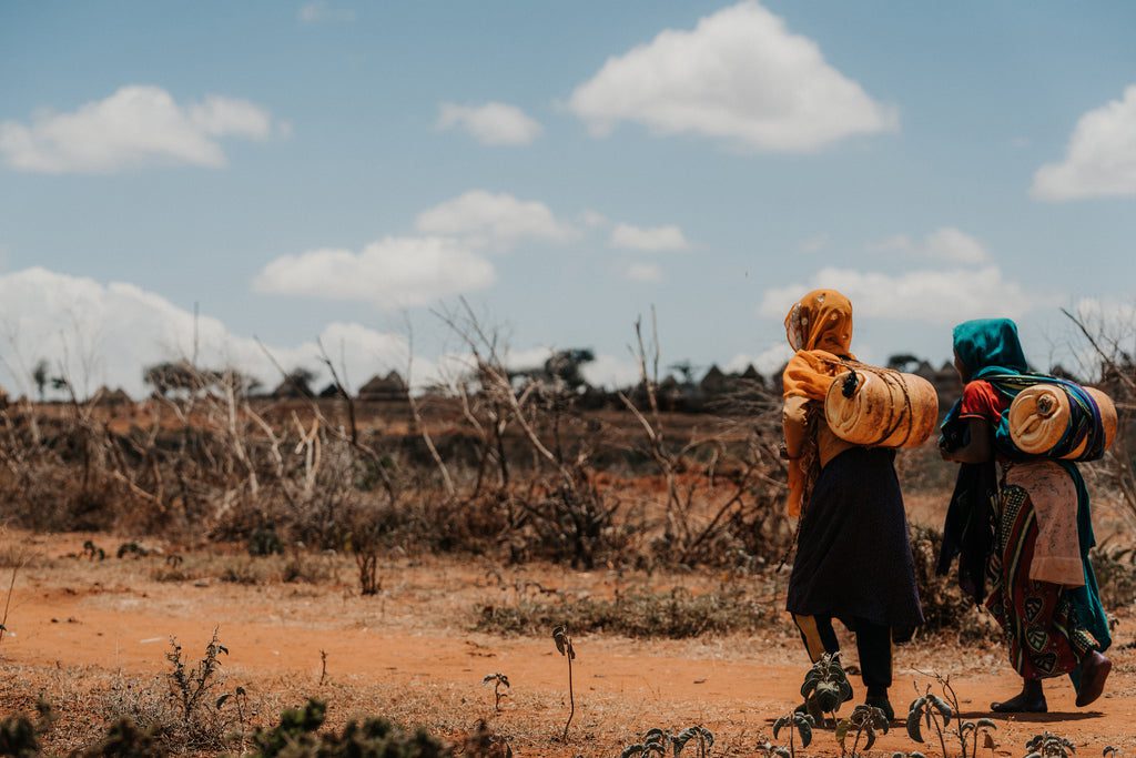 Two women and a child walk across a dry, barren landscape under a partly cloudy sky, carrying rolled-up blankets or mats on their backs. Sparse vegetation and leafless shrubs surround them.