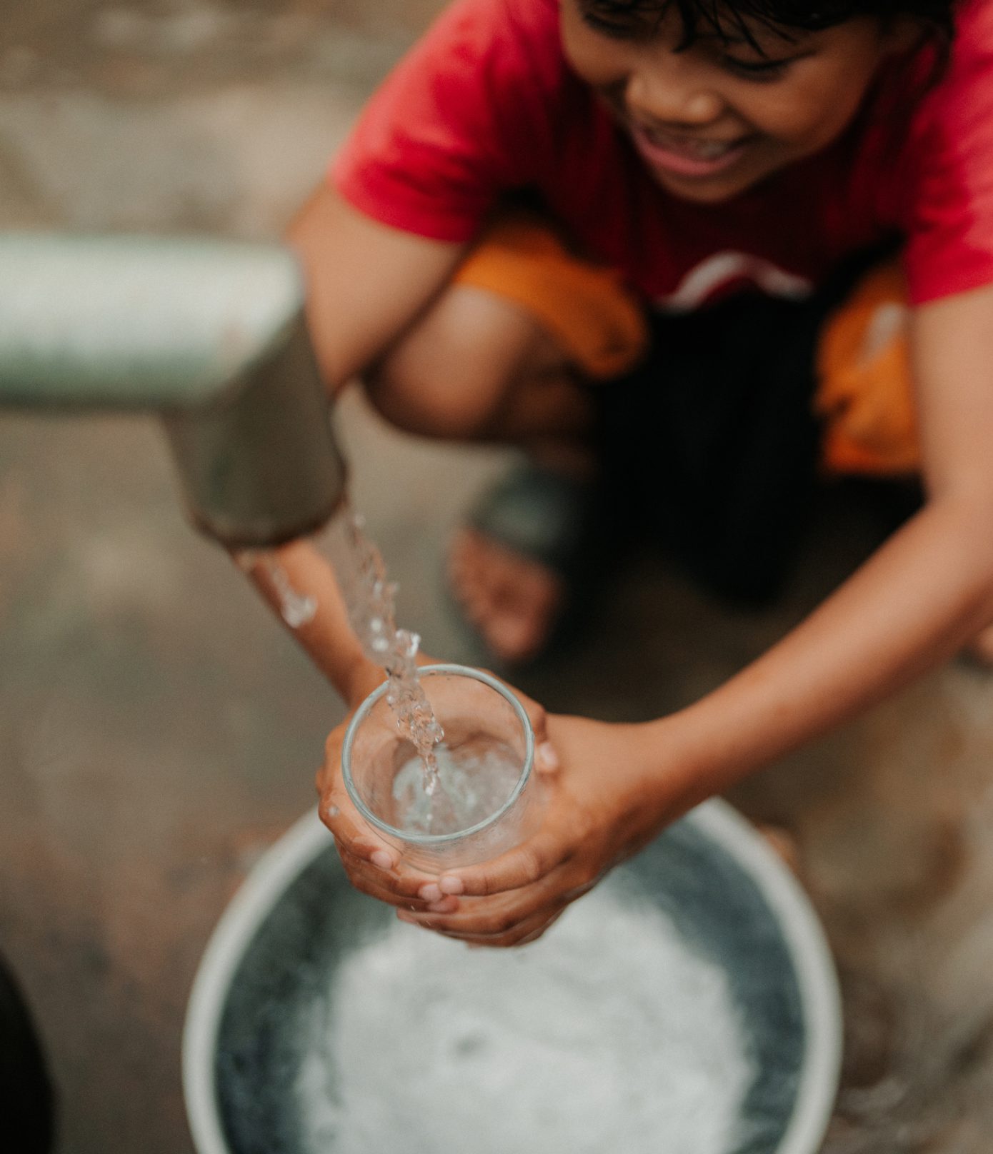 A smiling child in a red shirt fills a glass with water from an outdoor tap, holding the glass over a metal basin. The scene is viewed from above.