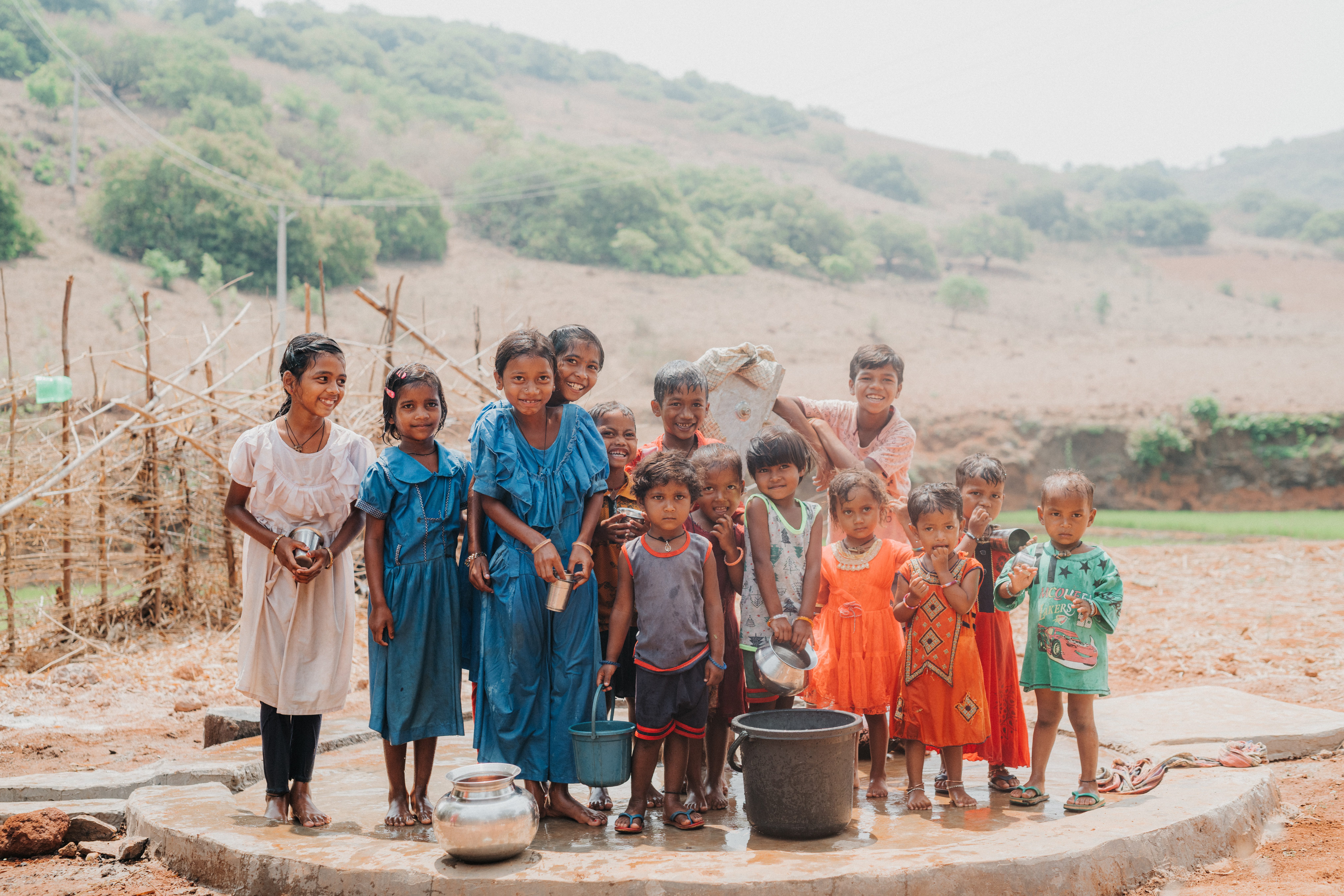 A group of children in colorful clothes stand together outdoors near a water pump, smiling and holding metal containers, with hills and greenery in the background.