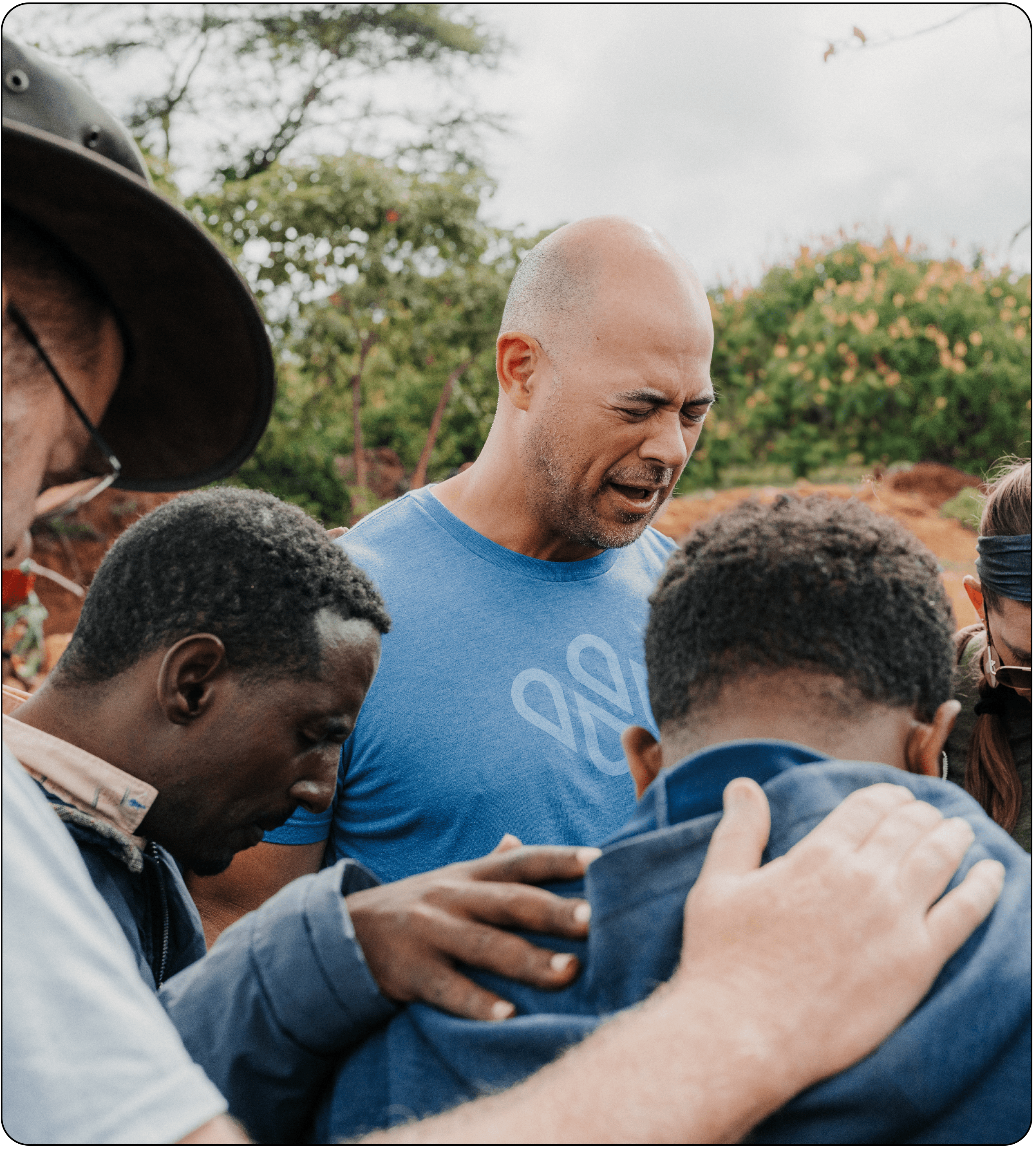 A group of people stand close together outdoors, some with their arms on each others shoulders. One man in a blue shirt appears to be leading the group in prayer or reflection. Trees and greenery are in the background.