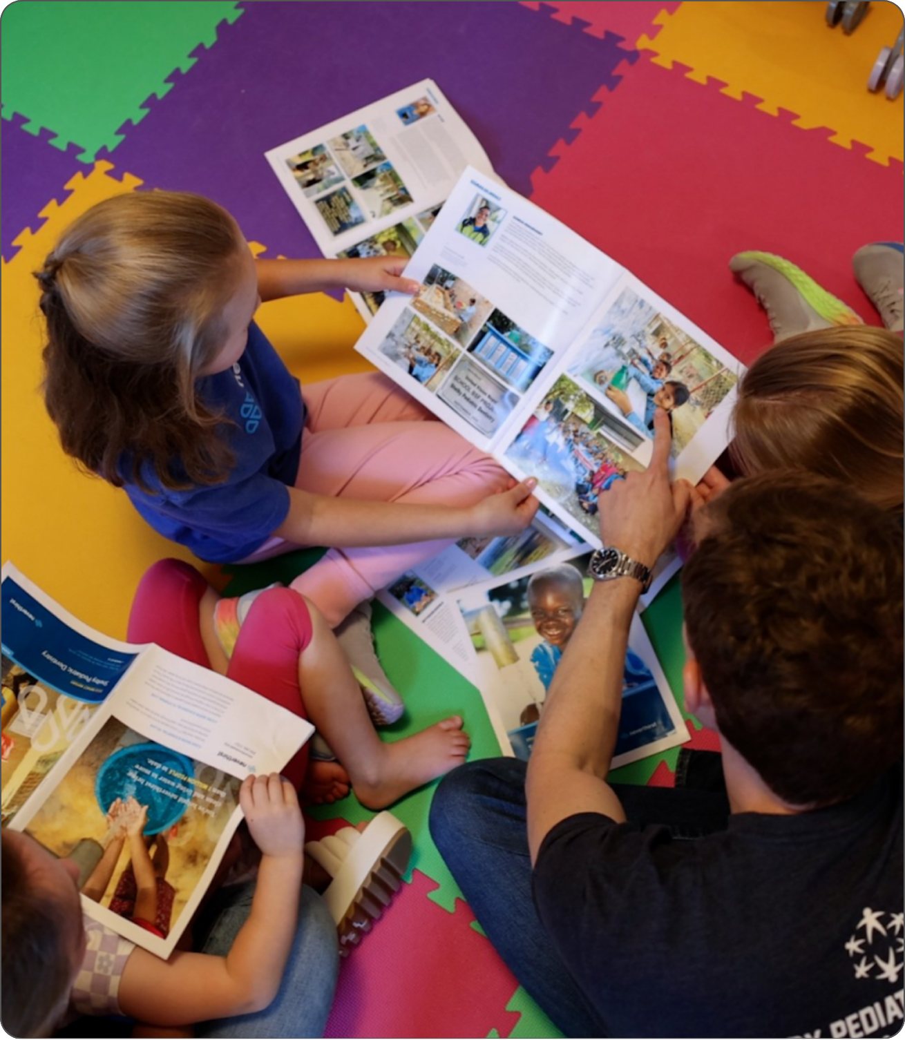 Four people, including two children and two adults, sit on a colorful foam mat, looking at and discussing open books or magazines with photos and text.