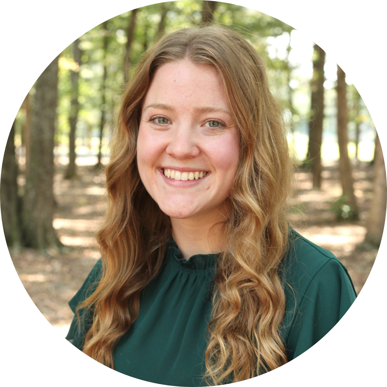 A woman with long wavy light brown hair, wearing a dark green top, smiles while standing outdoors in a sunlit forest with tall trees in the background.