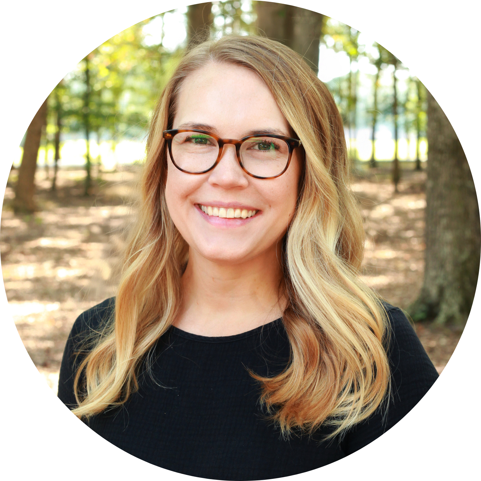 A woman with long blonde hair and glasses smiles while standing outside in a sunlit forest. She is wearing a black top and the background shows trees and sunlight filtering through the leaves.