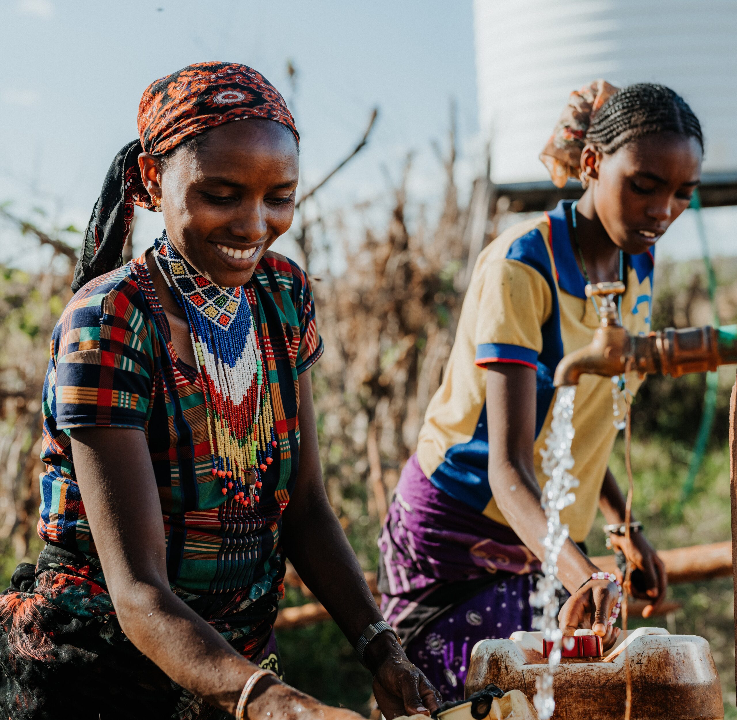Two women in colorful traditional clothing and beaded jewelry fill containers with water from a communal tap outdoors, smiling and working together. A large water tank is visible in the background.