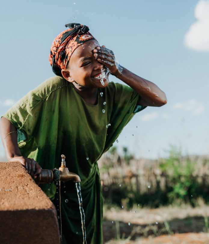 A smiling child in a green shirt splashes water on their face from an outdoor tap, with water droplets visible in the air and greenery in the background under a clear sky.