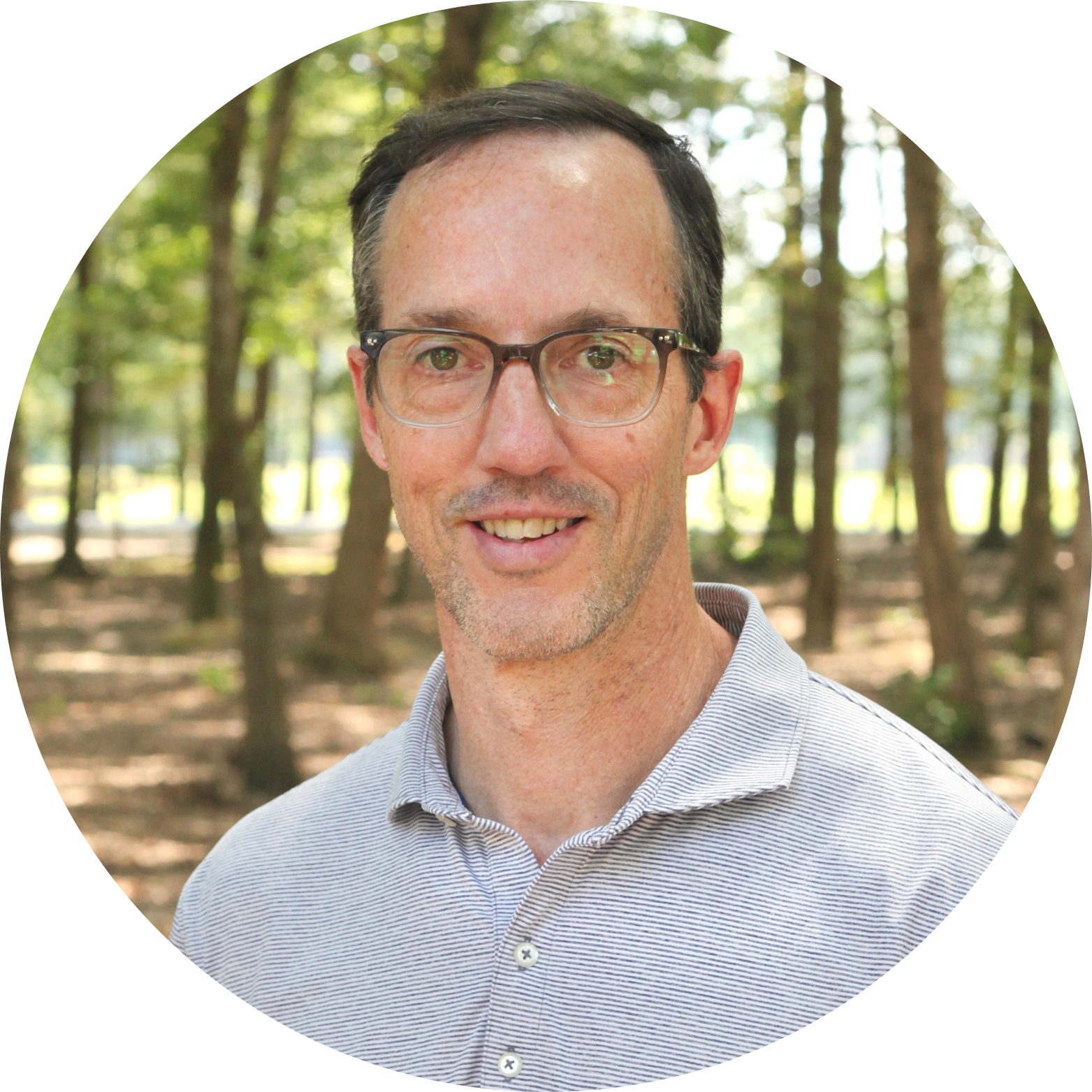 A man with short brown hair and glasses, wearing a light gray striped polo shirt, stands outdoors in a sunlit forest with trees in the background. He is smiling and looking at the camera.