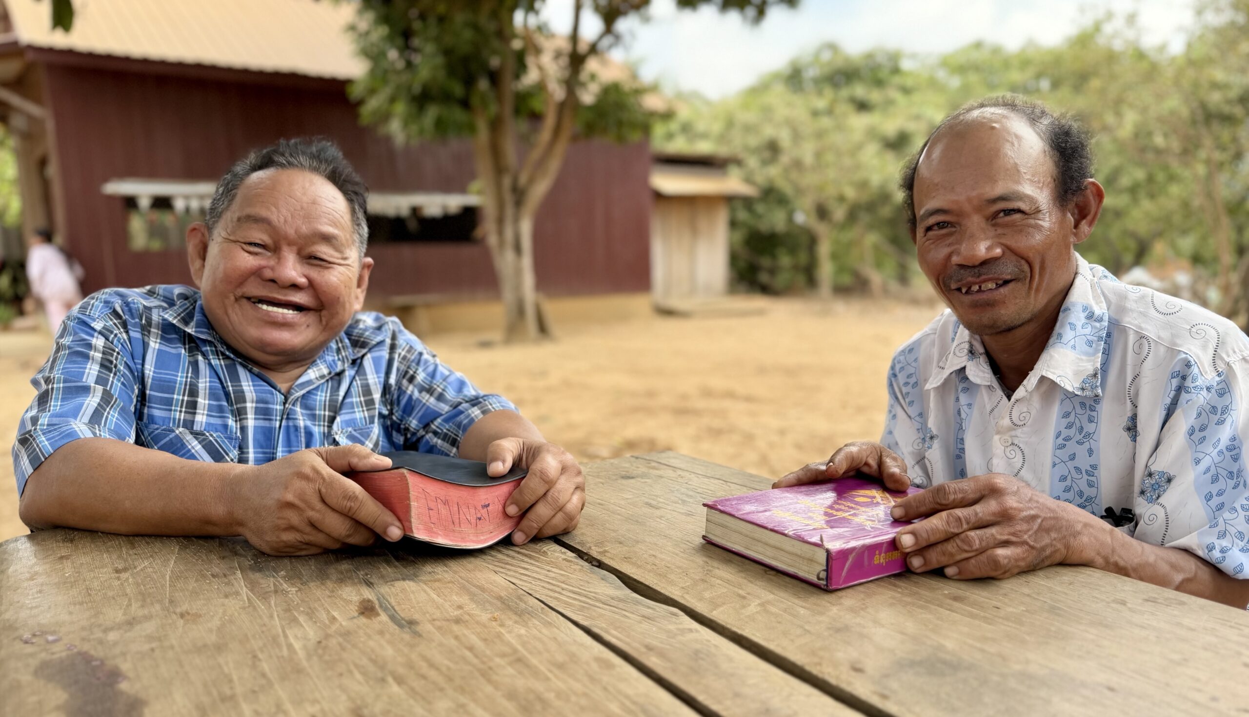 Two men sit at a wooden table outdoors, smiling and holding books. Trees and wooden buildings are visible in the background on a sunny day.